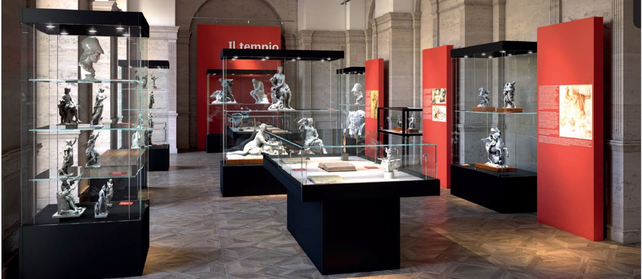 A museum exhibition room with glass display cases containing sculptures and artifacts. Red walls feature informational panels, and the floor has a geometric pattern. The space is well-lit and modern, with classical architectural details.